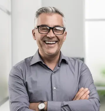Smiling man with gray hair and glasses, wearing a gray shirt and watch, with arms crossed.