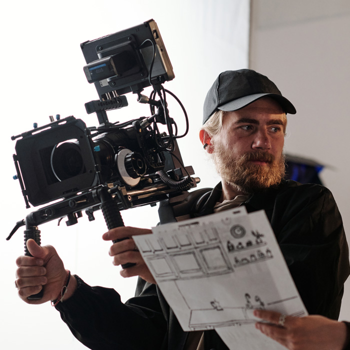 Man holding a camera setup with one hand and a script in the other, wearing a black cap and jacket.