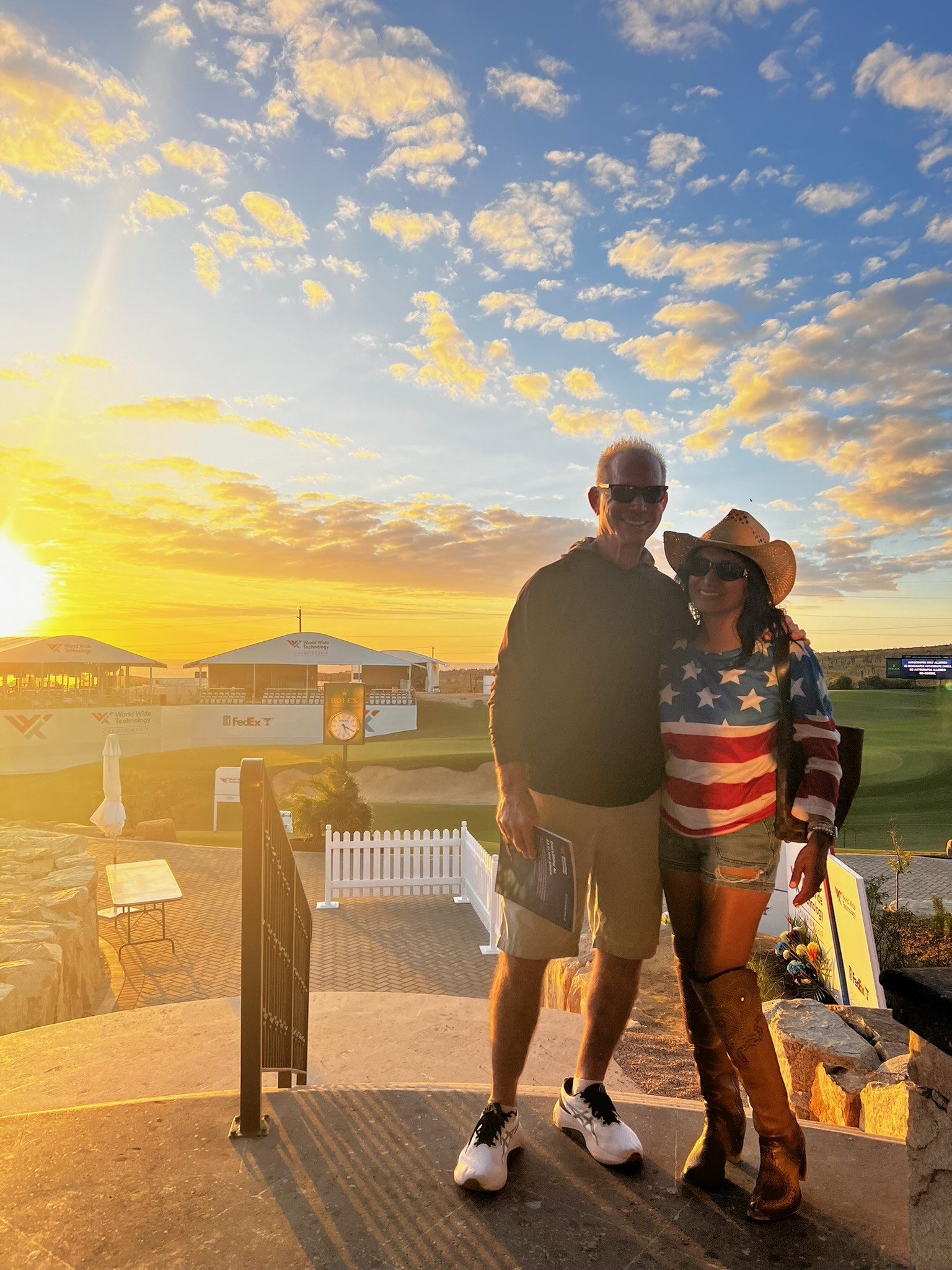 Two people posing for a photo at sunset on a golf course, with one wearing an American flag shirt and cowboy hat.